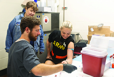 JoEllen Sefton looks on as Cole Bordonie conducts a lab blood draw on a member of the military.