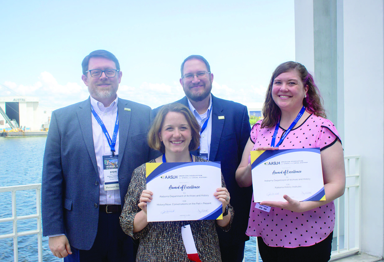 Two men and two women standing on a balcony overlooking waterfront with women holding Award of Excellence certificates.