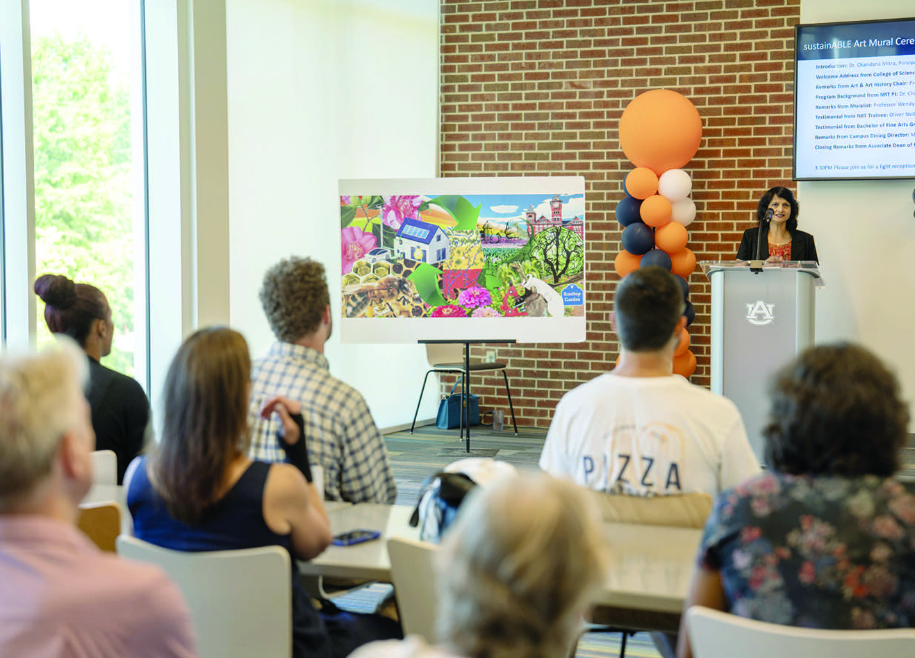 Woman speaker at a podium displaying mural to a group of onlookers