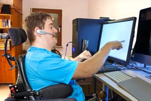 Young man in a wheelchair with physical disabled hands using a touchscreen computer, headset, and other adaptive technology.