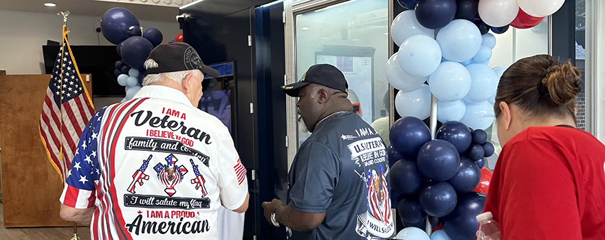 Two veterans standing inside the Chambers County Community Health and Wellness Center, with an OnMed Care Station visible in the background adorned with patriotic Veterans Day decorations, including flags and banners.