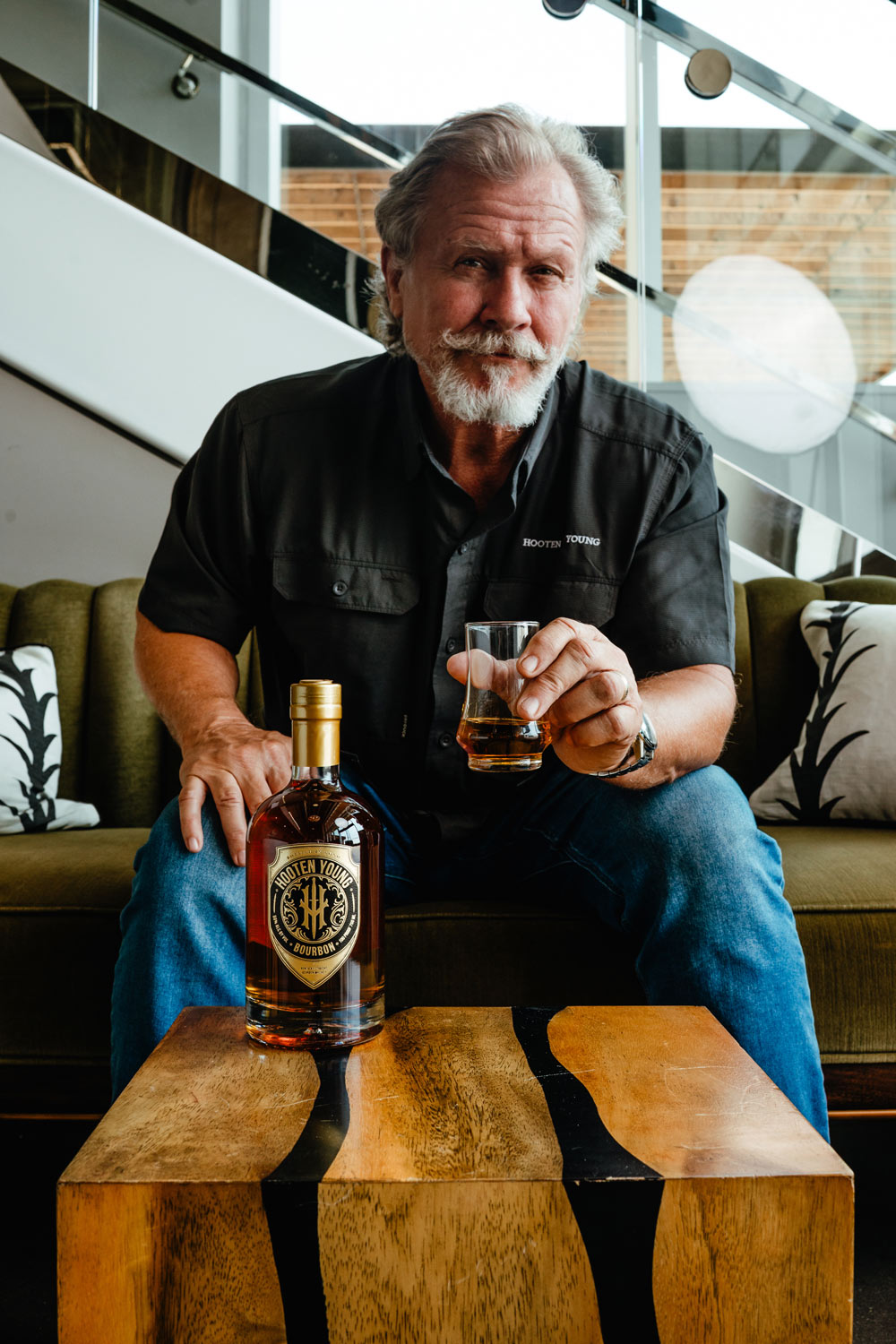 Photo of Norm Hooten holding a high ball glass with a bottle of whiskey in the front.