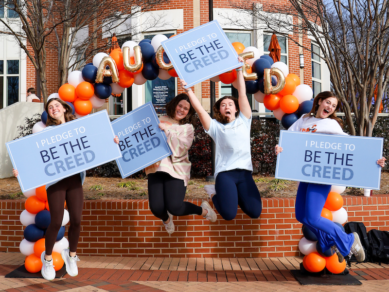 four students jumping into the air in front of a balloon arch at the student center holding signs that say I Pledge to be the Creed.